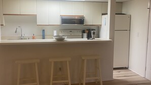 Een keuken met witte kastjes, een magnetron, een koelkast en twee barkrukken.