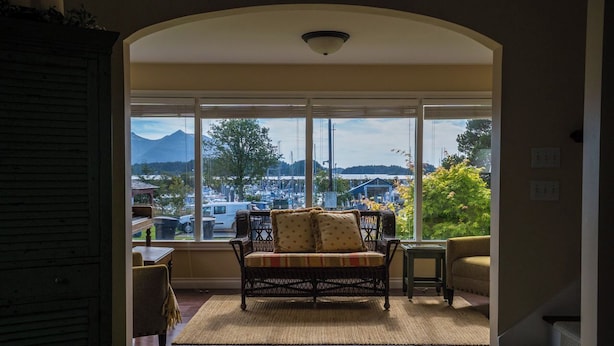 A cozy room with a wicker sofa and armchairs, a large window offering a view of a marina and mountains, and a small table with a lamp.