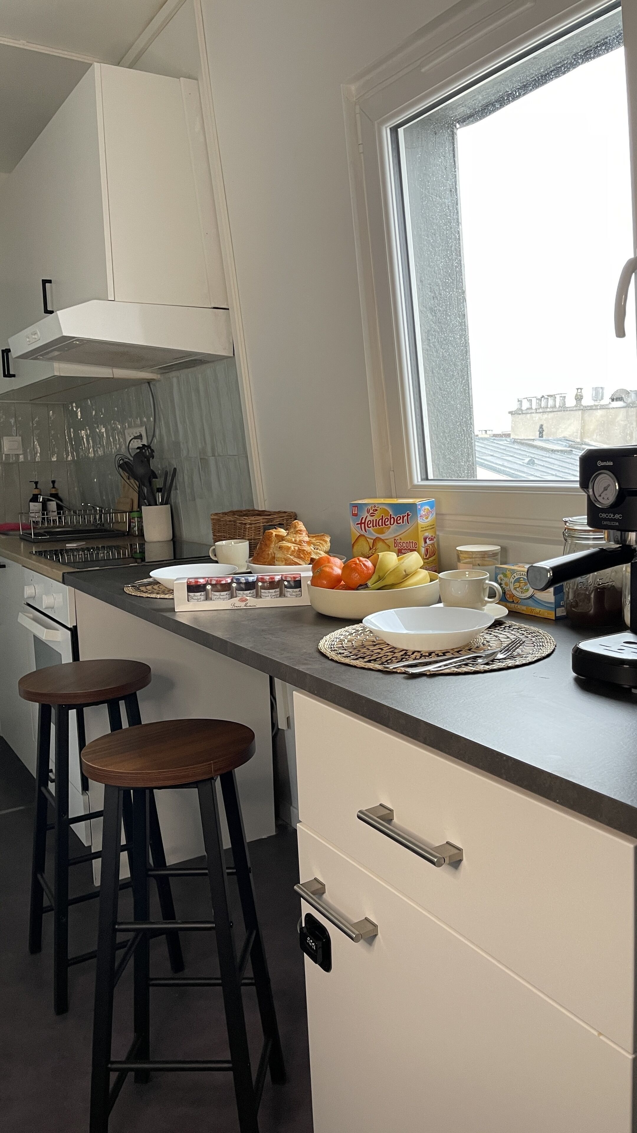Une cuisine moderne avec des meubles blancs, une grande fenêtre et un coin repas comprenant du pain, des fruits et une machine à café.