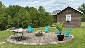 A wooden cabin with a circular seating area featuring a hammock, several blue Adirondack chairs, and a small fire pit. - The Cozy Den (Wellington)