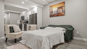 Un dormitorio con una cama, una silla, un cuadro y una kitchenette al fondo.
