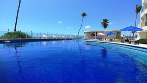 A swimming pool with lounge chairs and umbrellas, surrounded by palm trees and a clear sky. - Vista Mar, Piscina e Lazer no Areias do Mar (Barra de São Miguel)