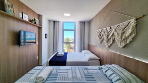 A hotel room with a bed, a TV, a window with curtains, and a macrame wall hanging. - Vista Mar, Piscina e Lazer no Areias do Mar (Barra de São Miguel)