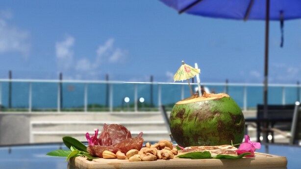A tropical drink setup with a coconut, snacks, and a decorative umbrella on a table. - Vista Mar, Piscina e Lazer no Areias do Mar (Barra de São Miguel)