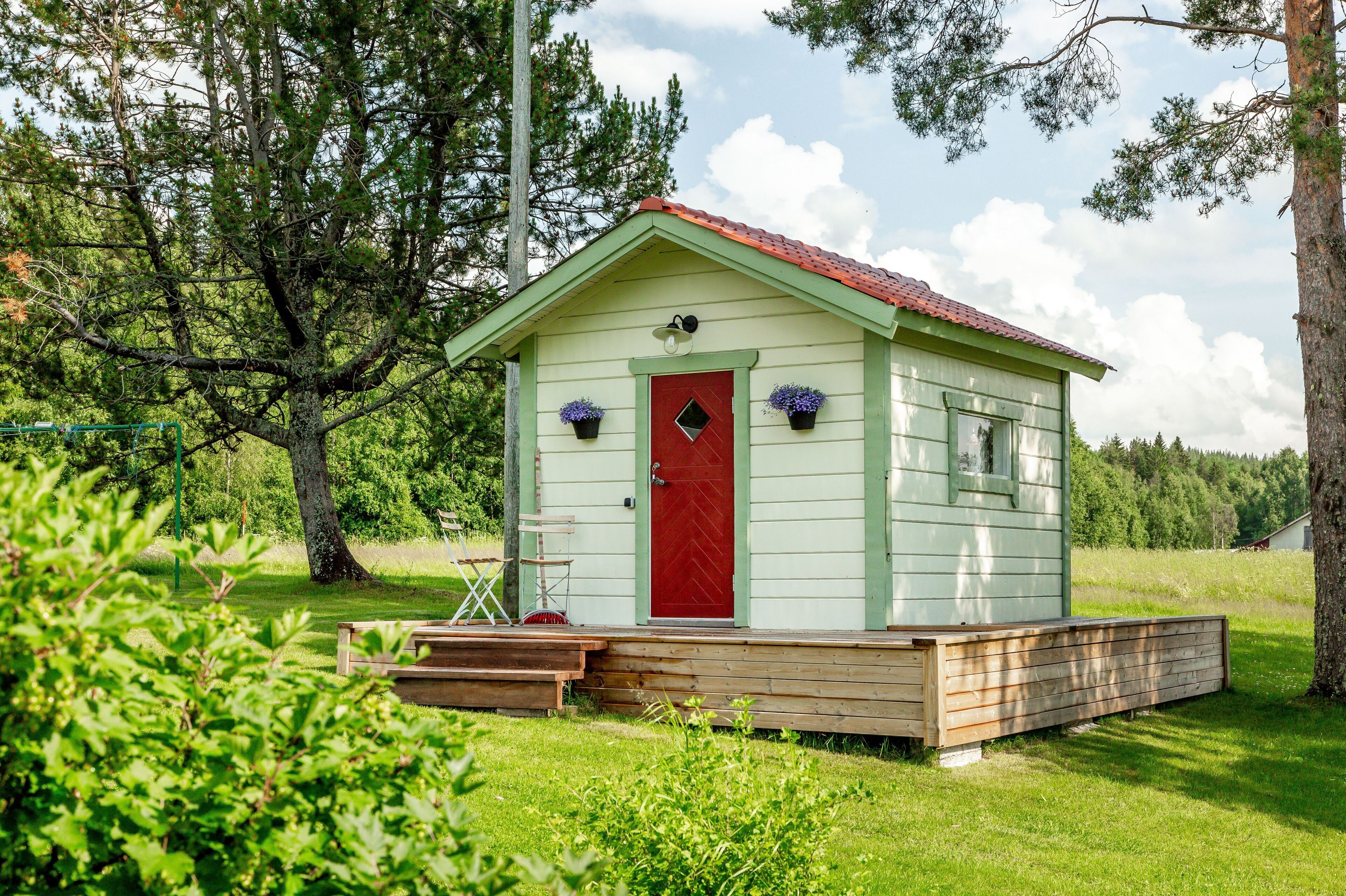 A small, single-story wooden cabin with a red door and a green roof, surrounded by trees and grass.