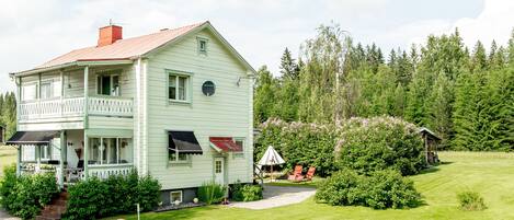 A two-story house with a balcony, surrounded by a well-maintained lawn and dense forest.