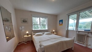 A bedroom with a bed, two bedside tables with lamps, a standing fan, a radiator, and a window with a view of trees. - Forest Family Home (Ytterhogdal)