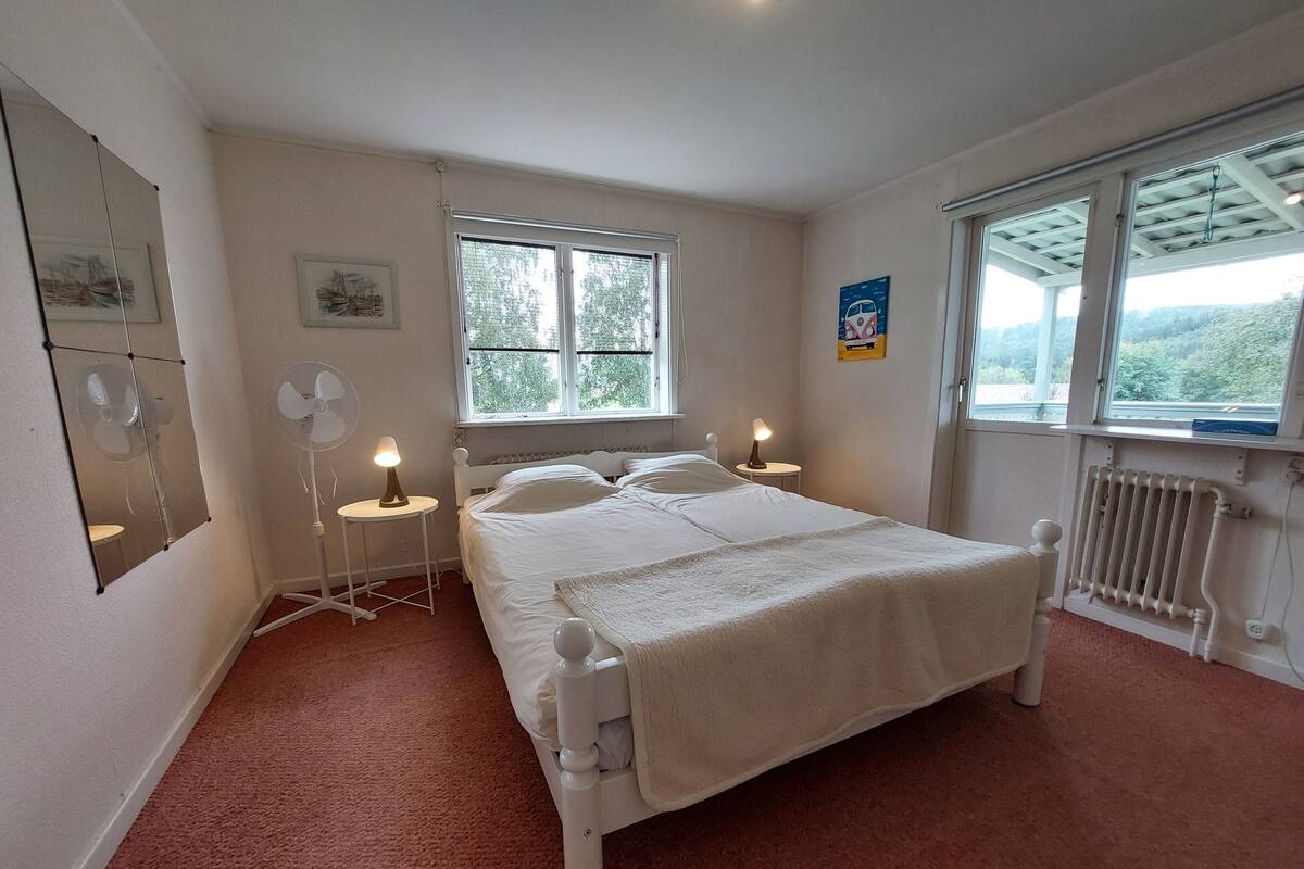 A bedroom with a bed, two bedside tables with lamps, a standing fan, a radiator, and a window with a view of trees.