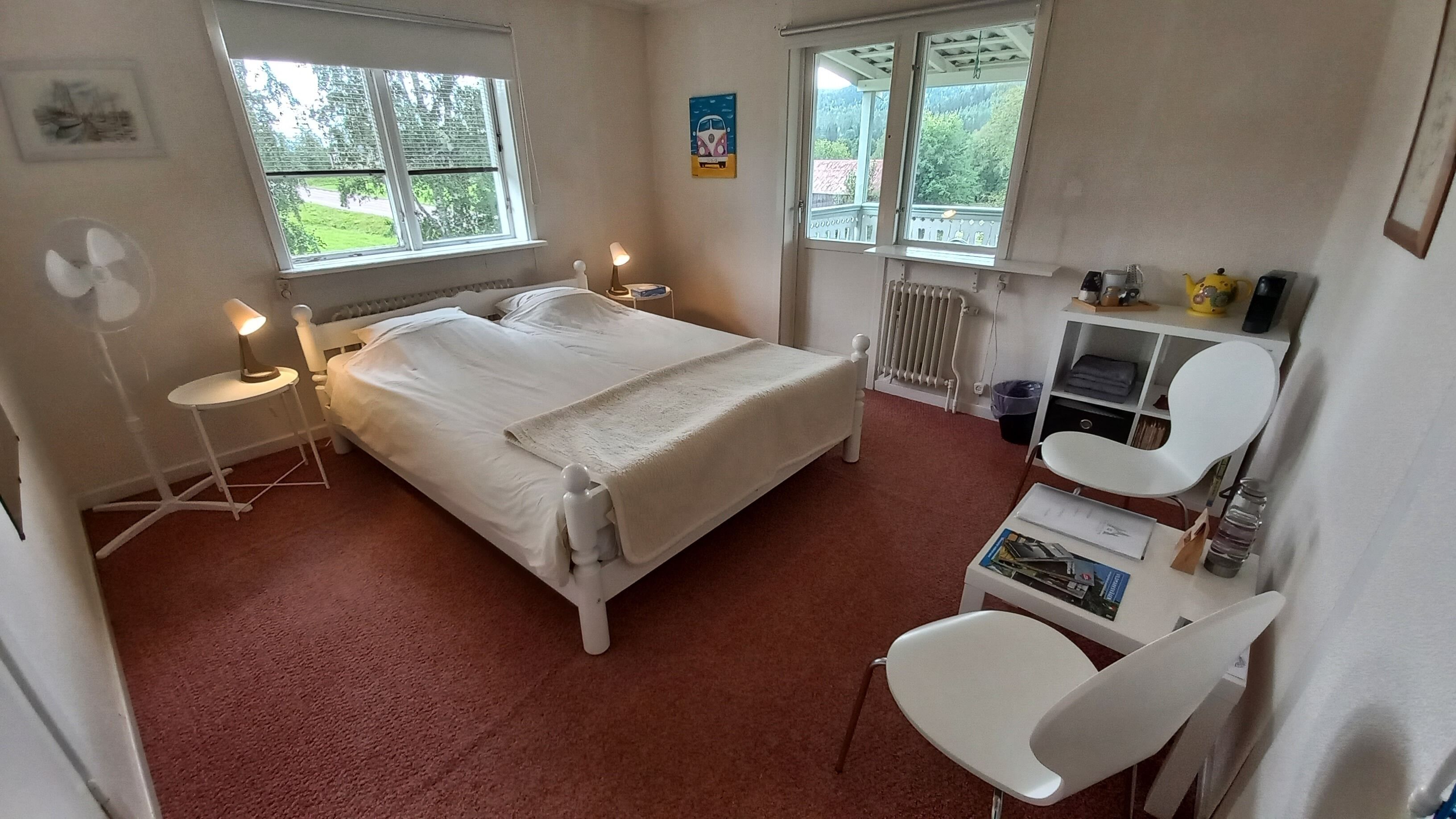 Une chambre à coucher comprenant un lit, des lampes de chevet, un radiateur, un bureau avec une chaise et des fenêtres donnant sur un paysage verdoyant.
