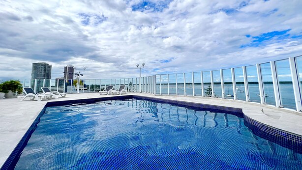 A rooftop swimming pool with a glass balustrade, lounge chairs, and a view of the city skyline and water. - Ocean View 2BR Apartment w/hot tub. Heated pool, Games room, Gym (Biggera Waters)
