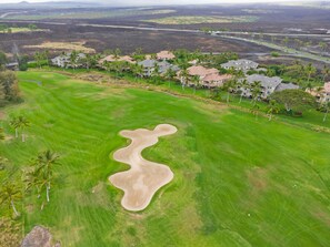 Um campo de golfe com uma zona de areia, palmeiras e edifícios residenciais ao fundo.