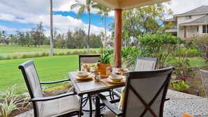 Une terrasse extérieure avec une table dressée pour le repas, entourée de chaises, et donnant sur un jardin luxuriant avec des palmiers et une vue sur un quartier résidentiel.