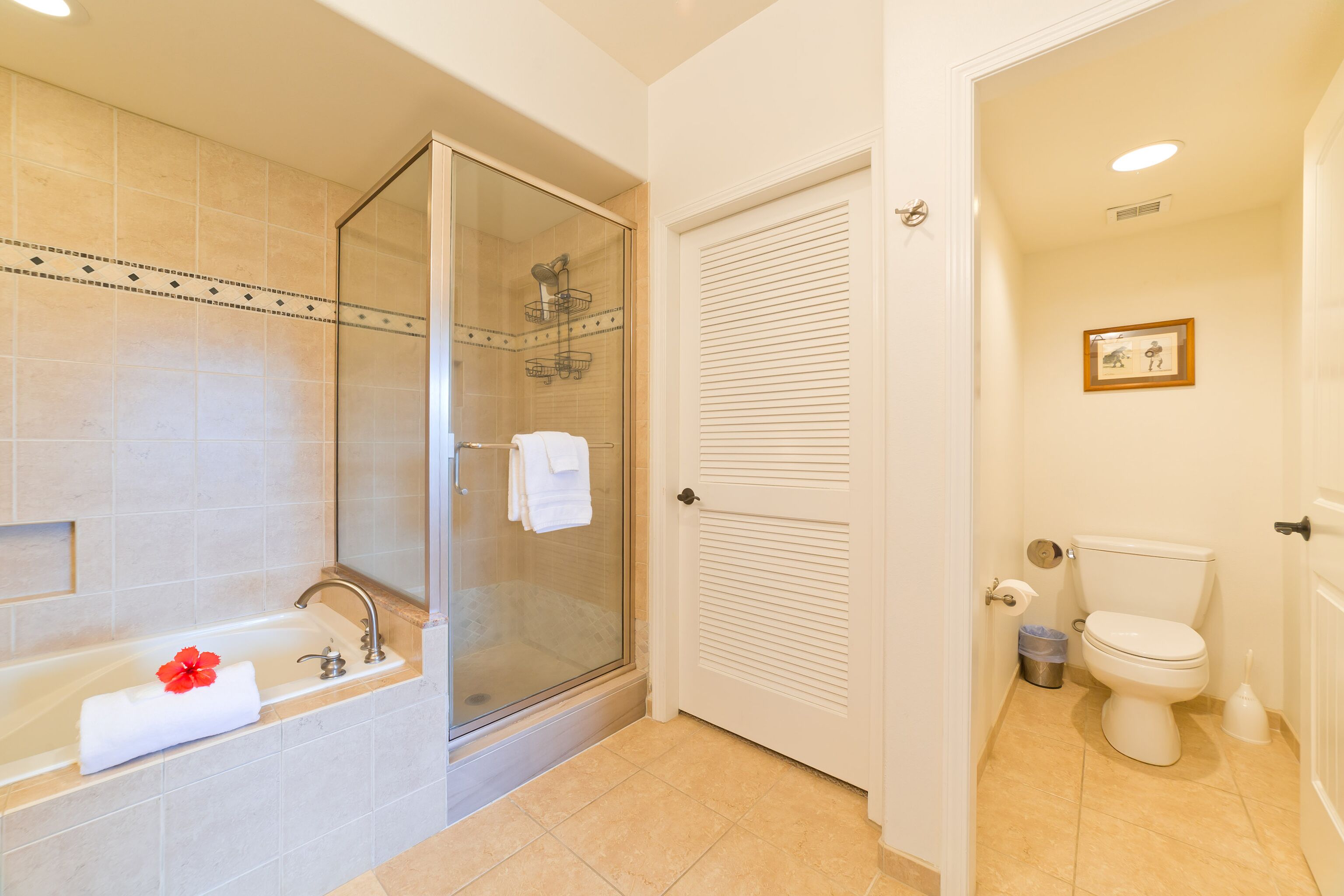 A bathroom with a glass-enclosed shower, a bathtub with a red flower, a toilet, and a framed picture on the wall.
