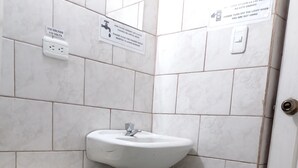 Ein Badezimmer mit Toilette, Waschbecken und Fliesenwänden. Schilder weisen auf Steckdosen hin und bitten darum, das Licht auszuschalten, wenn es nicht benötigt wird.