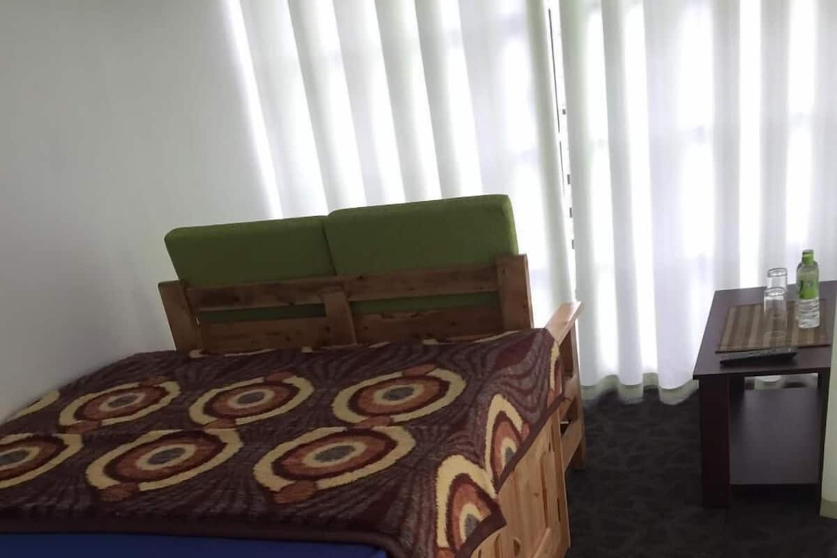 A room with a wooden bed, a small table with a bottle and glass, and a window with blinds.