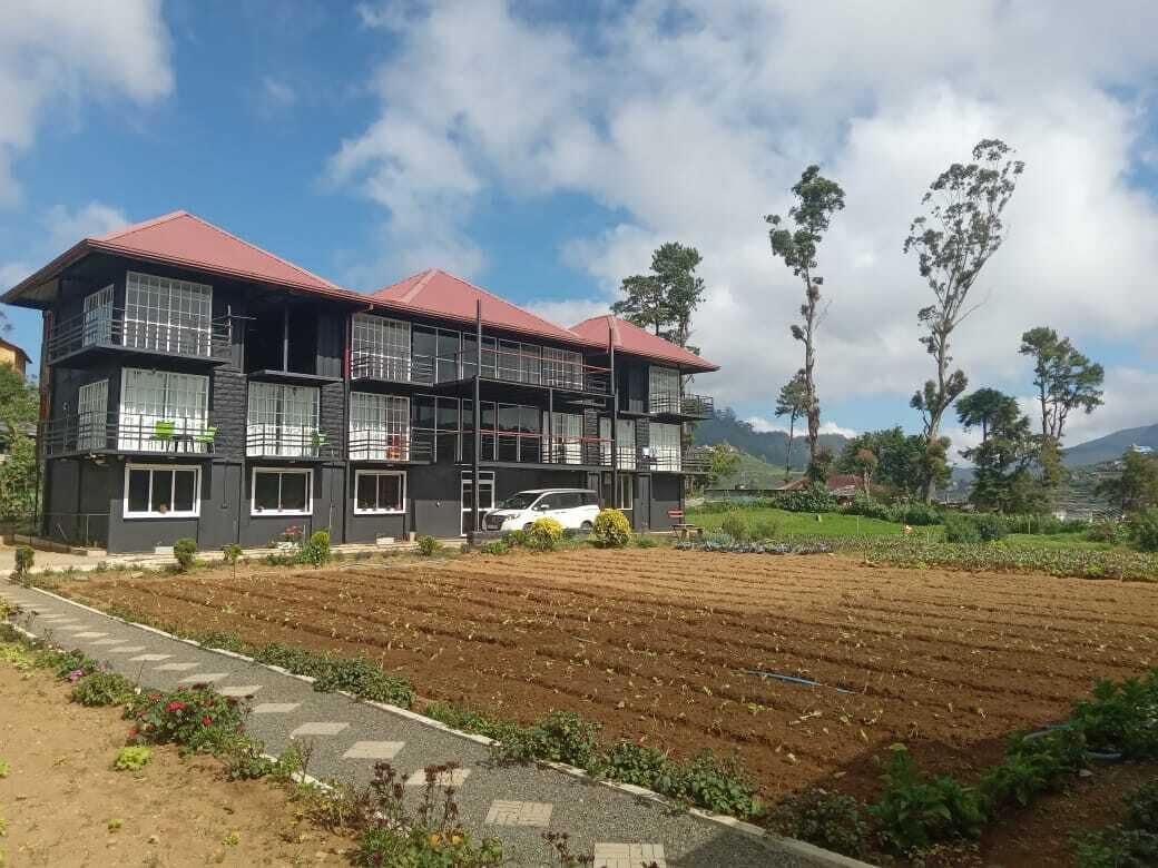 A modern two-story building with large windows and balconies, situated in a rural area with a field in the foreground and trees in the background.