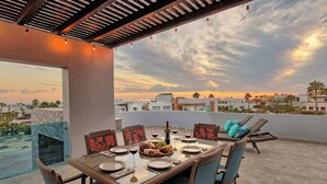 A rooftop terrace with a dining table set for four, chairs, a fireplace, and a view of the cityscape at sunset. - Modern Luxury Beachside Retreat (Puerto Peñasco)