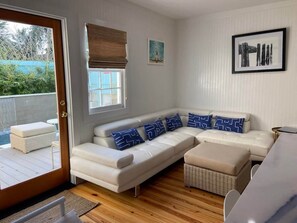 Relax in this serene space with a view of lush greenery, featuring a comfortable sofa, a framed art piece, and a wicker ottoman.