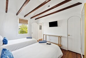 Two single beds with white bedding and blue pillows, a flat-screen TV, and a window with a view of greenery.