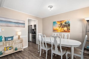 Un comedor con una mesa y sillas blancas, un cuadro de un atardecer con palmeras y una kitchenette al fondo.