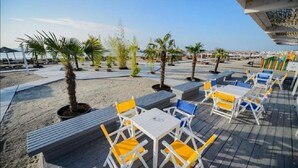 An outdoor seating area with tables and chairs, surrounded by potted plants and palm trees, overlooking a beach. - Alezzi Yaht Apartment With Sea View Near The Beach (MAMAIA SAT)