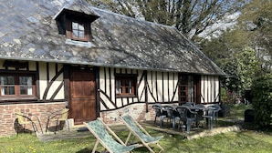 Ett traditionellt hus med korsvirkesfasad och sluttande tak, en trädörr och ett litet fönster. Det finns en trädgård med gräsmatta, en solstol och ett bord med stolar.