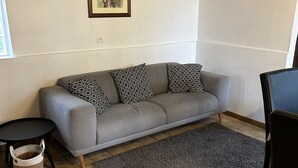 Ett vardagsrum med en grå soffa, mönstrade kuddar, en mörk matta, ett litet runt bord och en inramad tavla på väggen.