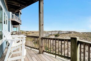 A wooden deck with white chairs, a wooden railing, and a view of the beach and dunes.