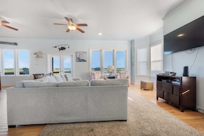A spacious living room with a grey sofa, a ceiling fan, and a flat-screen TV mounted on the wall. - Saagara Thira (Carolina Beach)