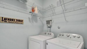 A laundry room with two white washers and dryers, a sign that reads 'Laundry Today or Naked Tomorrow,' and a metal shelf with laundry supplies.