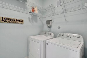A laundry room with two white washers and dryers, a sign that reads 'Laundry Today or Naked Tomorrow,' and a metal shelf with laundry supplies.