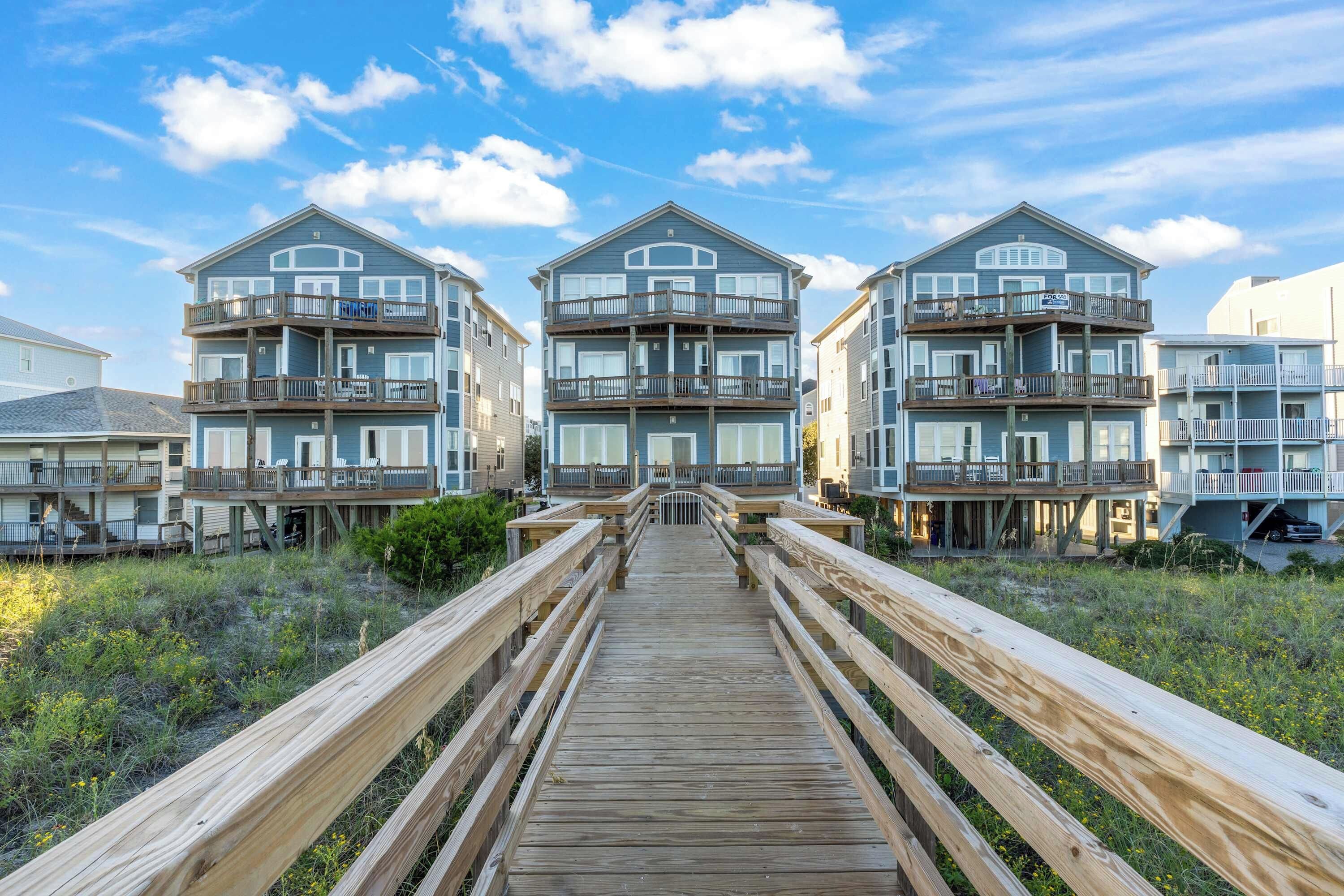 A wooden boardwalk leading to a beachfront property with multiple balconies and a clear sky.