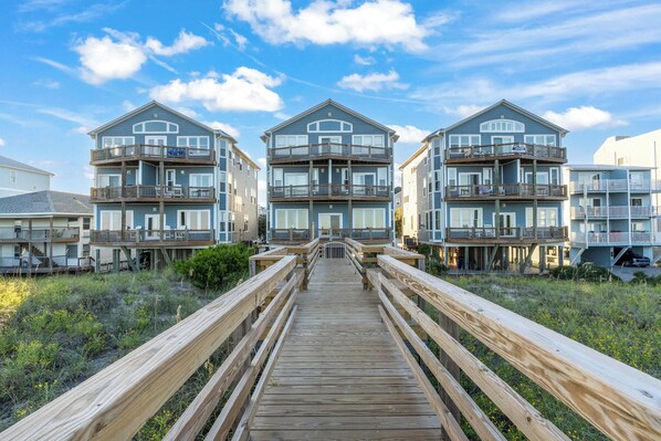A wooden boardwalk leading to a beachfront property with multiple balconies and a clear sky.