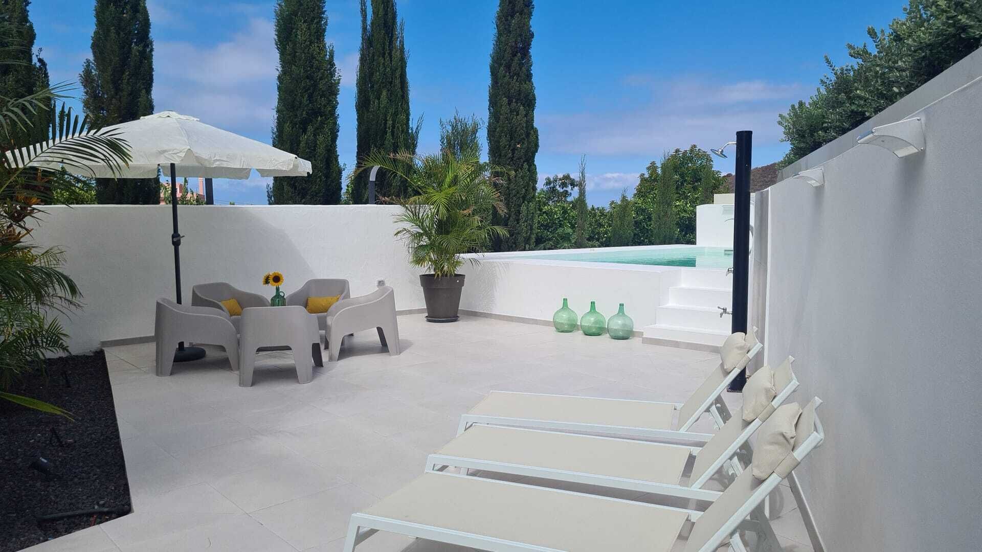 A rooftop terrace with white lounge chairs, a table with chairs, a potted plant, and a small pool.