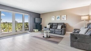 A modern living room with a grey sofa set, a flat-screen TV, and a balcony with a view of greenery. - Oceanfront Serene 2-bedroom 2 bath in Indian Shores Florida. (Indian Shores)