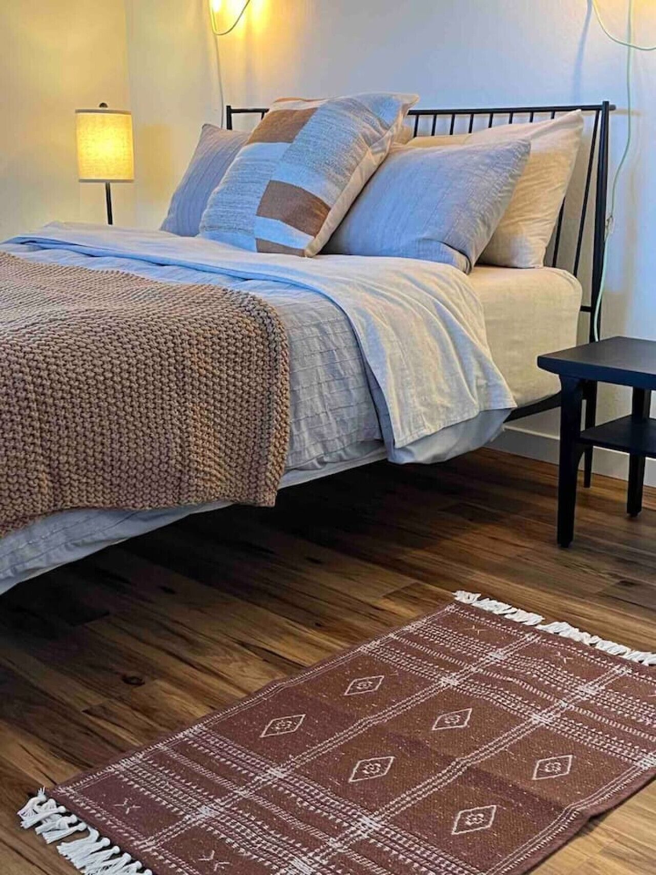 Una cama bien hecha con una colcha y almohadas estampadas, una mesita de noche pequeña y una alfombra estampada sobre un suelo de madera.