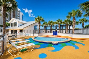 Zona de piscina con sillones de descanso, palmeras y un edificio con balcones.