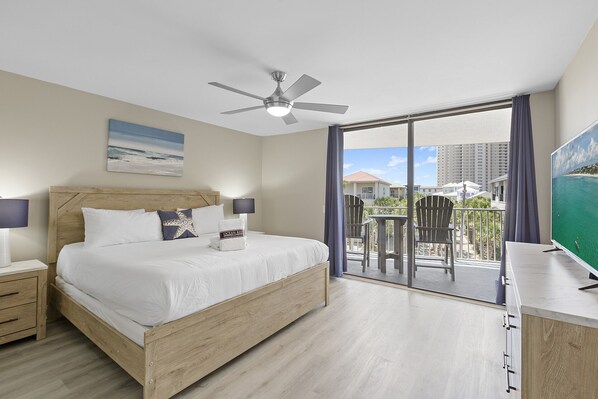 A hotel room with a bed, a ceiling fan, a TV, a balcony with chairs, and a view of the ocean and buildings.
