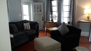 A living room with a dark-colored sofa, a striped ottoman, a small wooden table, and a lamp on a side table. - Cozy Private Home @ Blue Moon Cottage in Historic Deland, Fl. (DeLand)