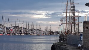 A marina with multiple sailboats docked, a large wheel structure, and a stone building with a flag. - Maison 2 Proche de la mer (Saint-Lormel)