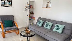 A living room with a grey sofa, a wooden rocking chair, a small round table with a decorative item, and a bookshelf with books and a small shelf with a figurine. - Maison 2 Proche de la mer (Saint-Lormel)