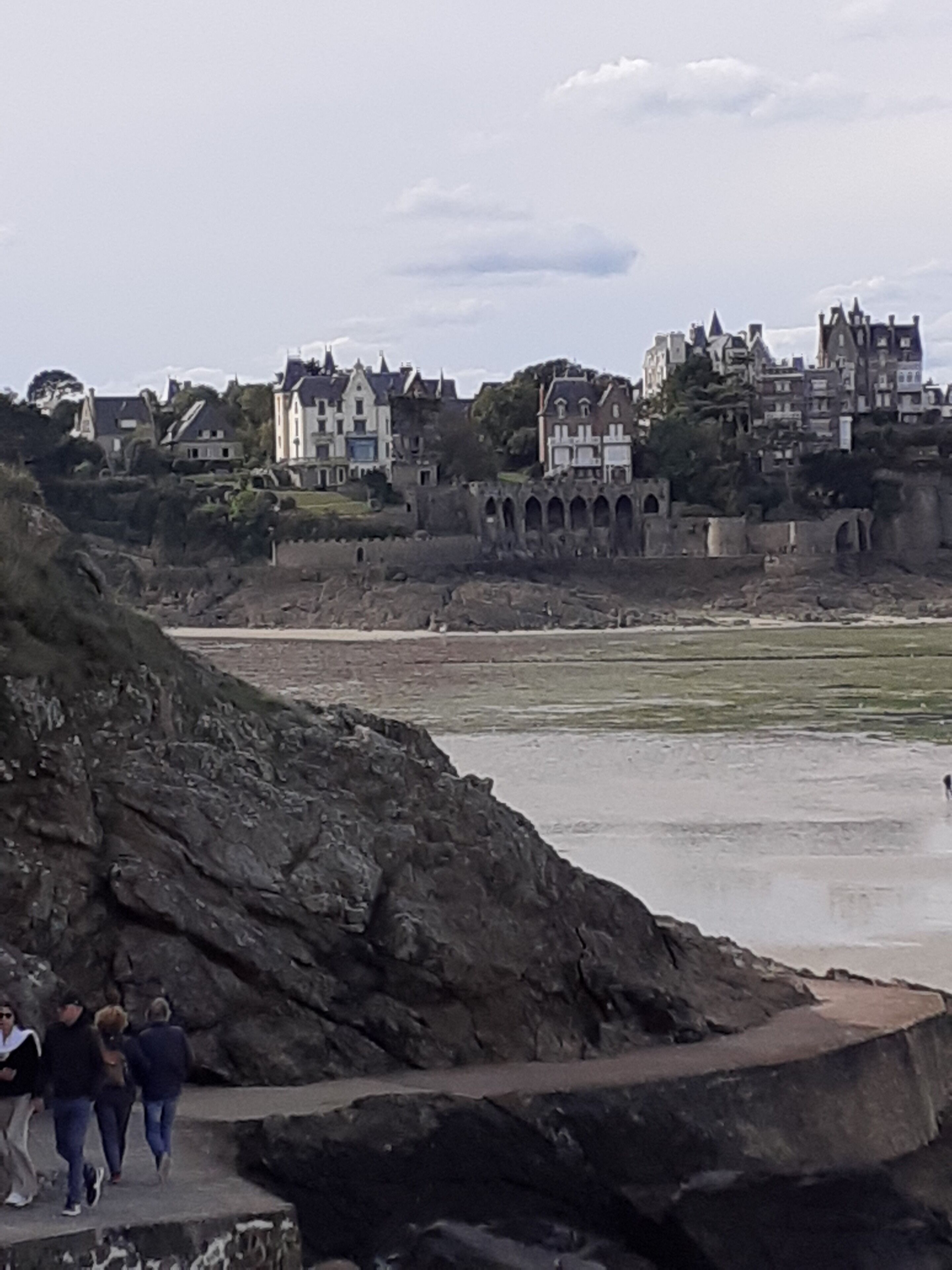 A coastal town with historic buildings, a rocky shoreline, and a paved walkway with people walking.