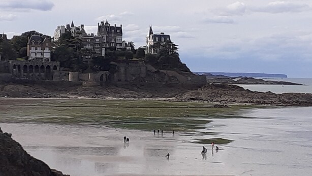 A coastal area with a castle-like building on a cliff, a beach with people, and a calm sea. - Maison 2 Proche de la mer (Saint-Lormel)