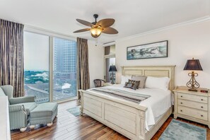 A hotel room with a large bed, a ceiling fan, a nightstand with a lamp, a chair, and a view of the city through a large window.