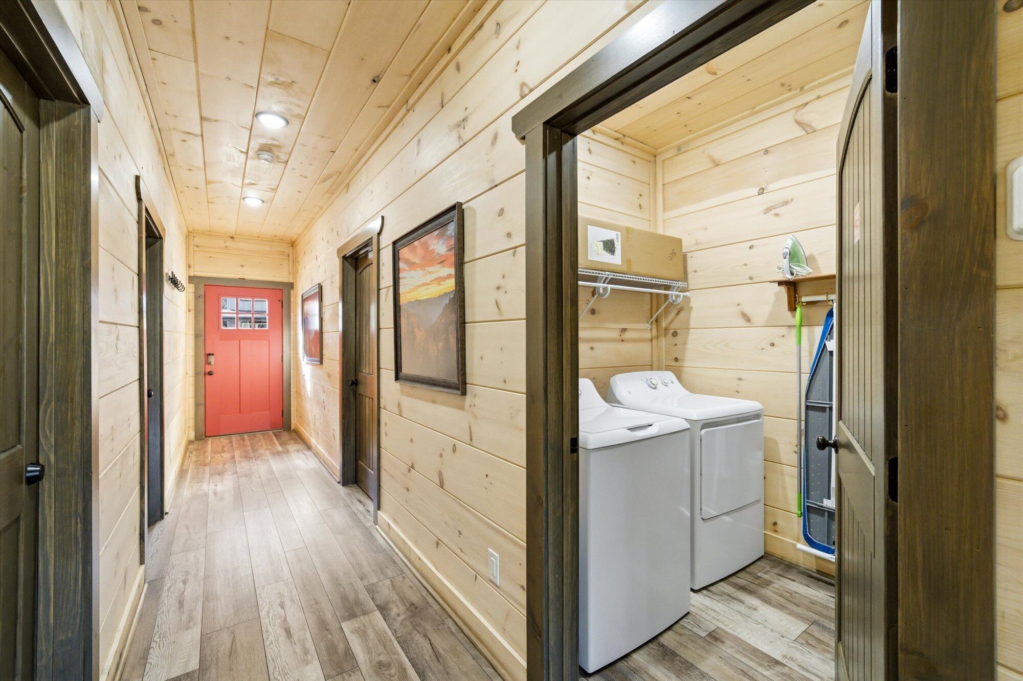 Un couloir étroit aux murs en bois, avec une porte rouge au fond. Dans un coin, on aperçoit deux appareils électroménagers blancs, qui pourraient être un lave-linge et un sèche-linge.