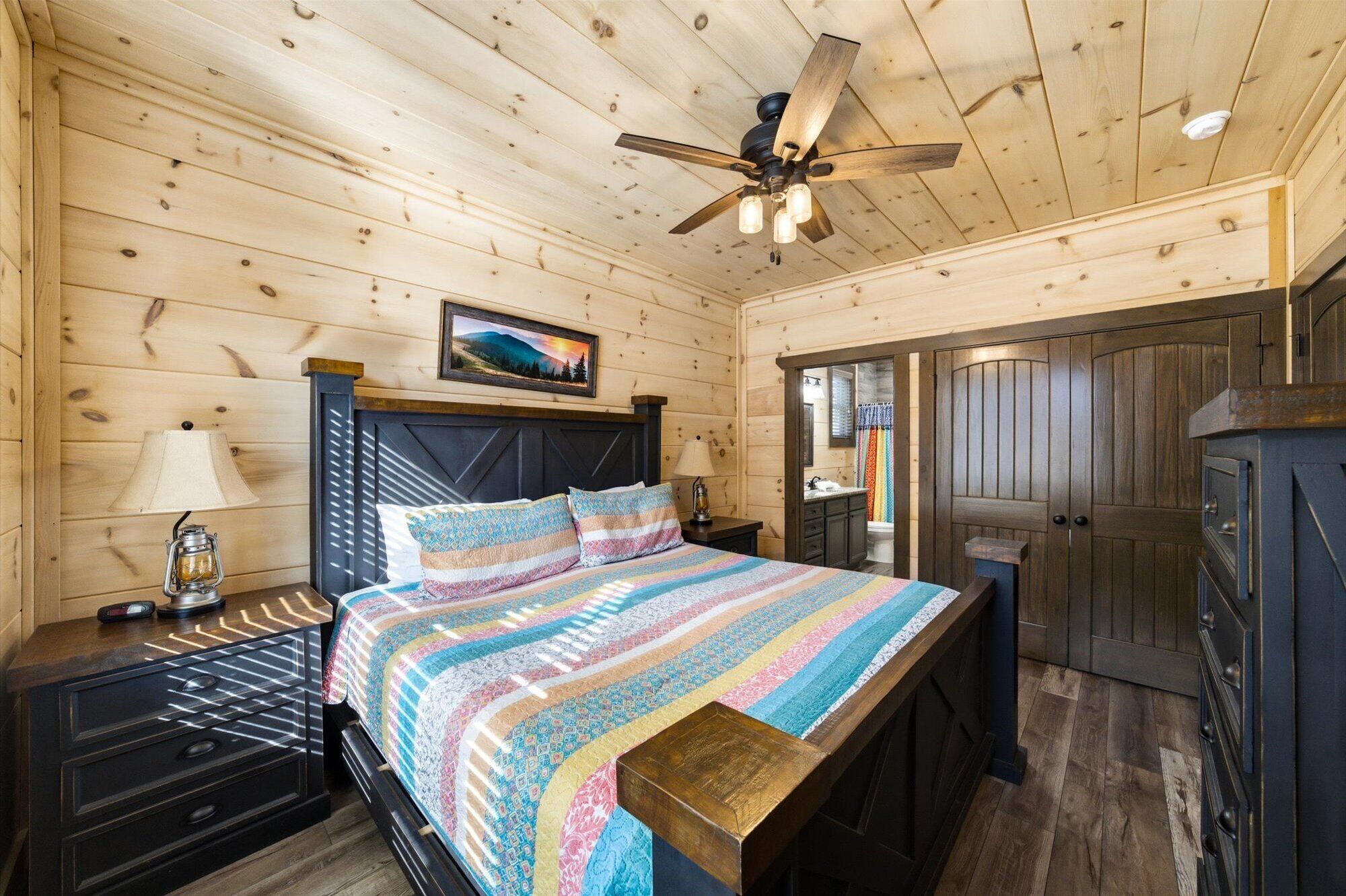 Un dormitorio de cabaña de madera con cama, mesita de noche, lámpara, ventilador de techo y un baño visible a través de una puerta abierta.