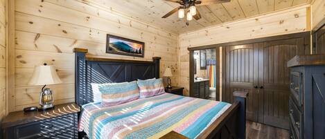 Un dormitorio de cabaña de madera con cama, mesita de noche, lámpara, ventilador de techo y un baño visible a través de una puerta abierta.