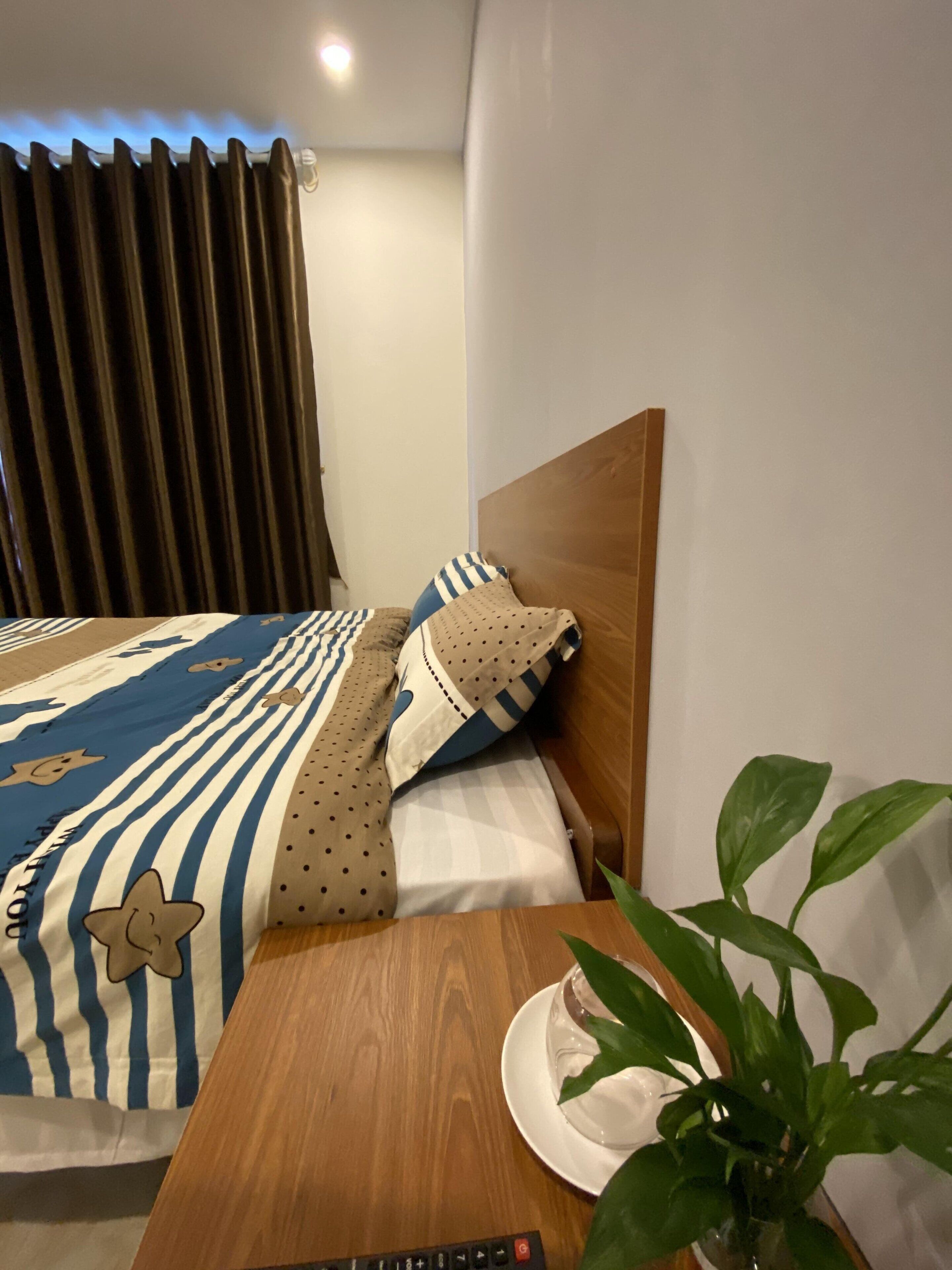 A neatly made bed with striped bedding, a wooden headboard, and a bedside table with a remote control, a cup, and a plant.