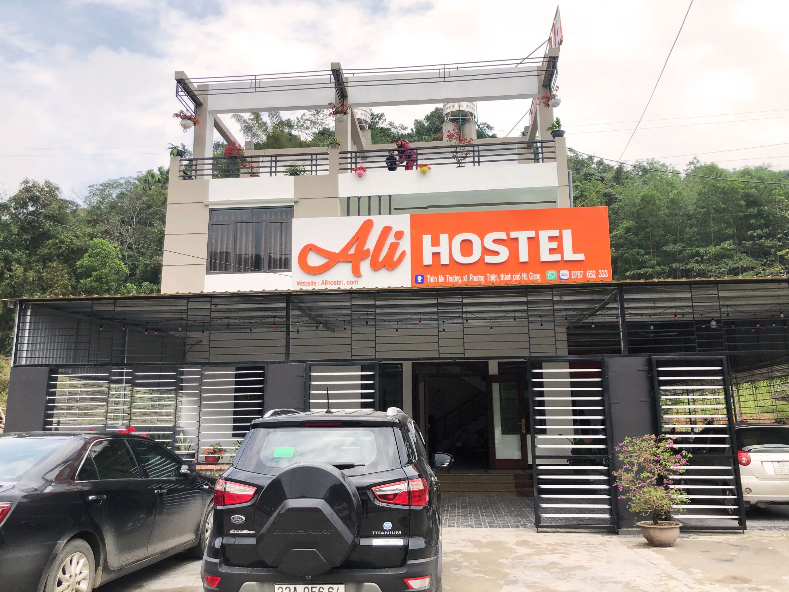A hostel with a prominent orange sign, a covered entrance, and parked cars.