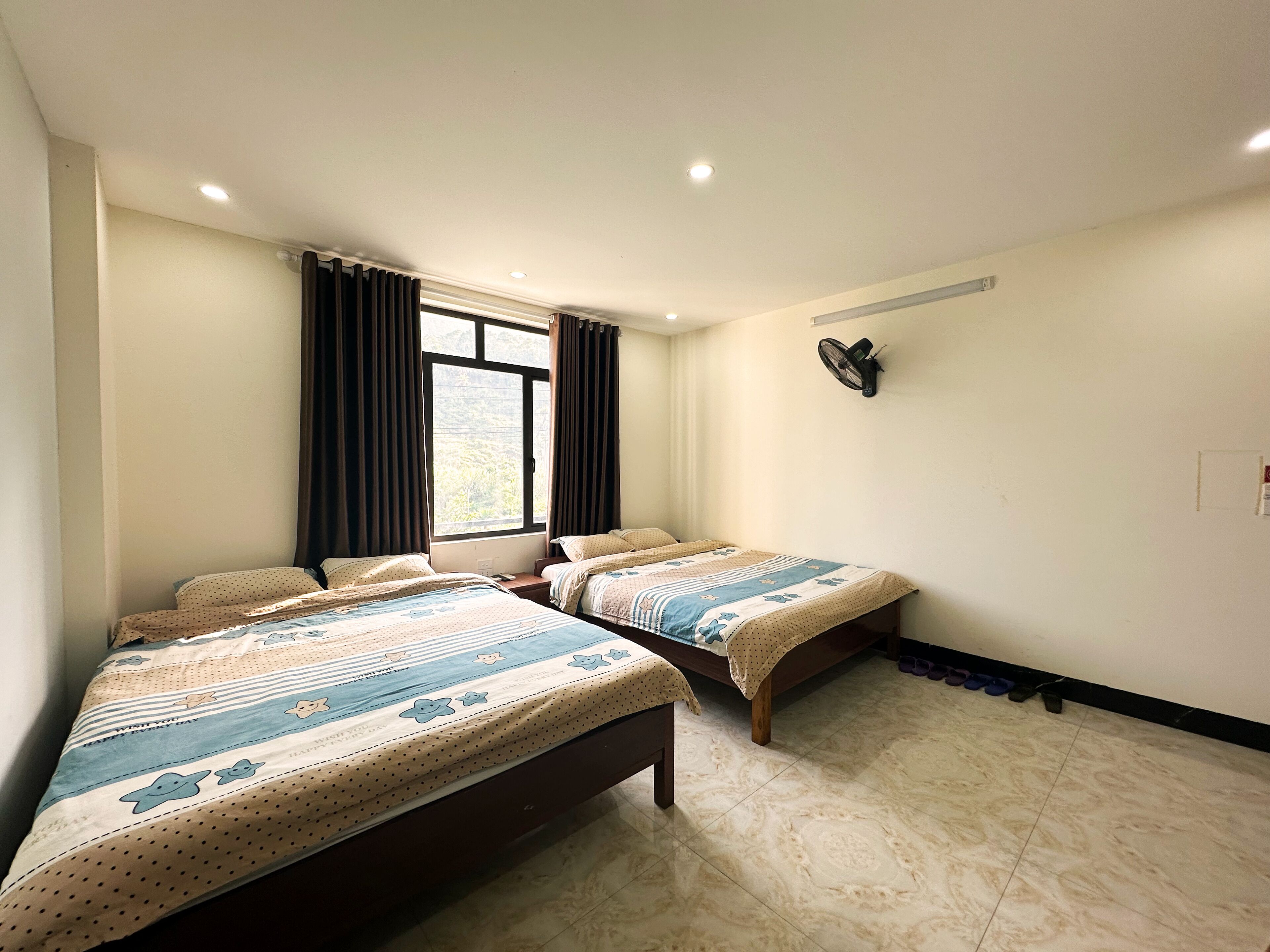 Une chambre d’hôtel avec deux lits, une fenêtre avec des rideaux, un ventilateur fixé au mur et un sol carrelé.